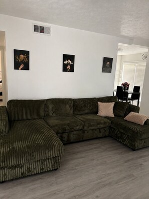 A sectional sofa with a textured fabric, a white wall with three framed artworks, and a dining area with a table and chairs.