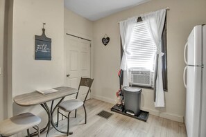A compact kitchen with a dining table, chairs, a refrigerator, a window with blinds, a wall-mounted heater, and a door. - Niagara Falls Retreat (Niagara Falls)