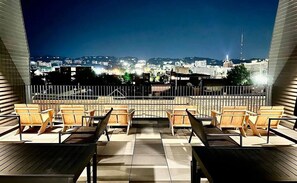 Terraço no telhado com cadeiras e mesas de madeira, proporcionando vista para a cidade à noite.