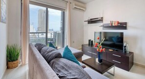 Ein modernes Wohnzimmer mit großem Fenster, einer Couch, einem Fernseher und einer dunklen Holzkommode.