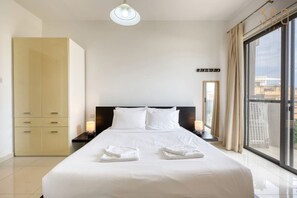 Ein Hotelzimmer mit einem großen Bett, einem Kleiderschrank, einem Nachttisch, einer Lampe und Blick durch ein großes Fenster.