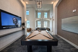 A pool table with a television displaying a sports event, a seating area with a view of the outdoors, and a large screen on the wall.