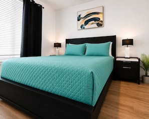 A bed with a quilted teal bedspread, a black headboard, two bedside tables with lamps, a wall-mounted abstract painting, and a potted plant.