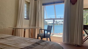 A bedroom with a bed, a chair, a table, and a large window overlooking a body of water. - Villa Martinique Canals (Jeffreys Bay)