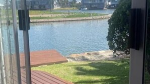 A view through a glass door to a deck with a water body, grass, and buildings in the background. - Villa Martinique Canals (Jeffreys Bay)
