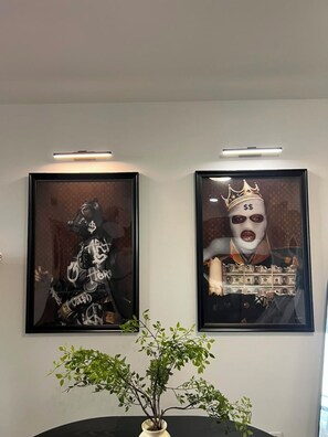 Twee ingekaderde kunstwerken aan een muur, een potplant op de voorgrond en een plafondlamp.