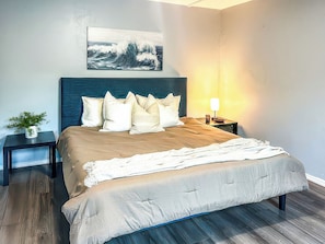 A bedroom with a bed, pillows, a bedside table with a lamp, a wall-mounted artwork, and a small plant. - Stylish Getaway (Bodfish)