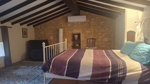 Ein Schlafzimmer mit Steinwand, einem Bett mit gestreifter Decke, einem Sessel, einem kleinen Tisch mit einer Vase und einem Fenster mit Bilderrahmen.
