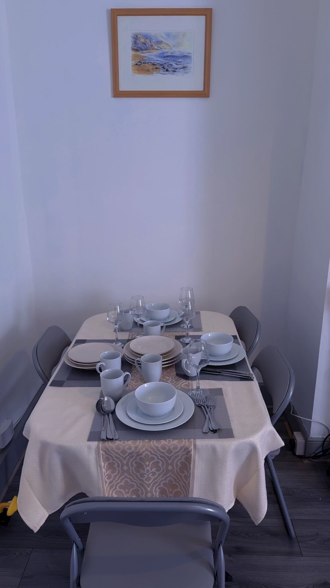 Una mesa de comedor dispuesta con platos, vasos, cubiertos, sillas, un cuadro en la pared y un mantel con motivos.