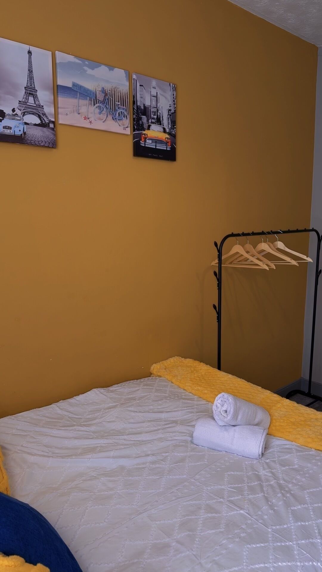 A hotel room with a bed, a towel, a coat rack, and three wall-mounted pictures.