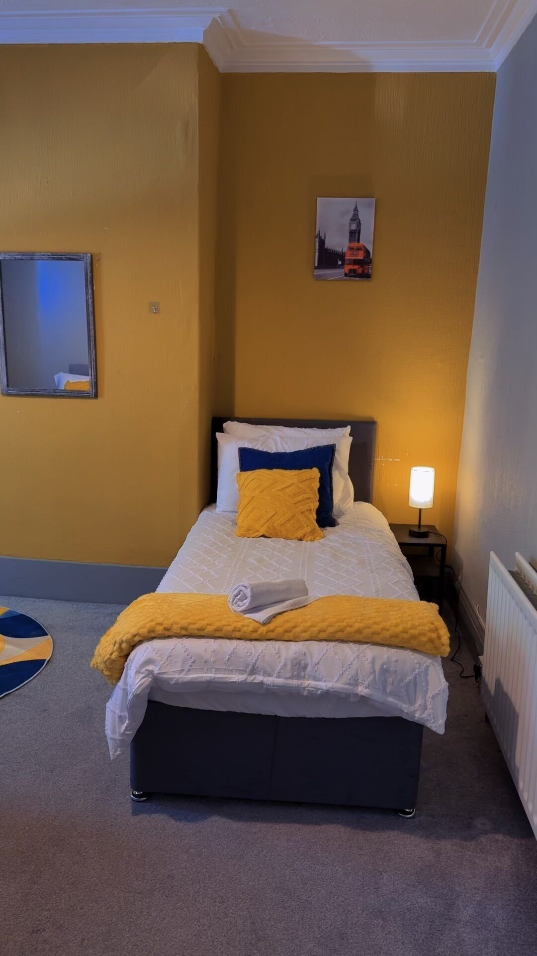 Un dormitorio con cama, mesita de noche, lámpara, un cuadro de un edificio y un espejo en la pared.