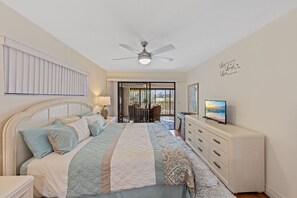 Ein Schlafzimmer mit einem Bett, einem Deckenventilator, einem Fernseher, einer Kommode und Blick durch eine Glastür.