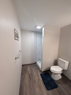 A bathroom with a toilet, a shower, a dark blue rug, and a picture on the wall.
