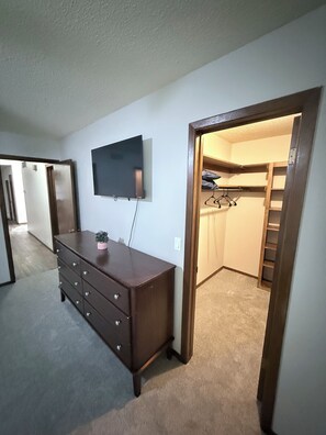 A room with a dresser, a television mounted on the wall, and a closet with a ladder.