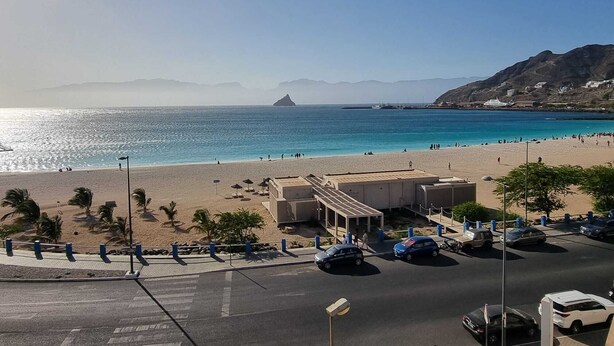 A beach with a parking area, a building with a covered section, and mountains in the background.