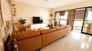 A modern living room with a leather sofa, a flat-screen TV, a wicker chair, a small table, and a large window with a view.