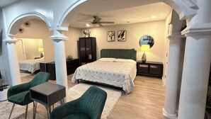 Quarto com cama, duas cadeiras verdes, mesa, espelho, ventilador de teto e mesa de cabeceira com abajur.