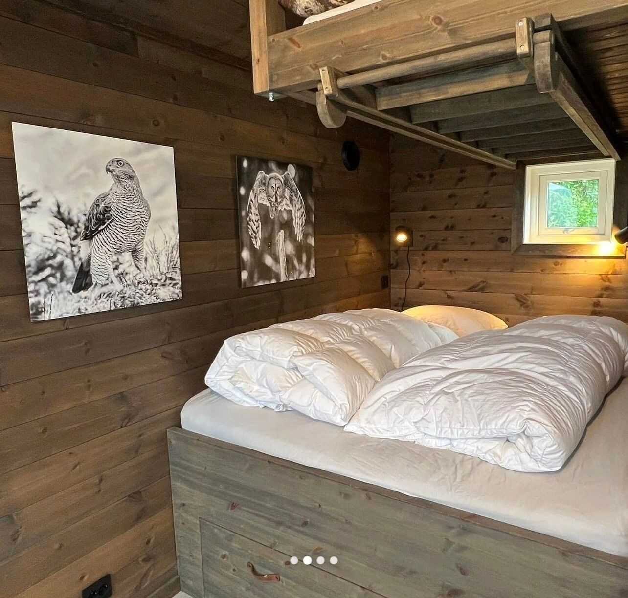 Ett sovrum i en stuga med träpanel, en säng, två konstverk som hänger på väggen och ett fönster med utsikt över grönskan.
