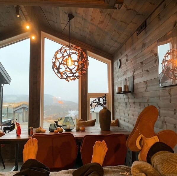 A cozy room with wooden walls, large windows, a dining table with chairs, and a hanging light fixture.