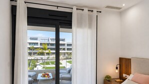Hotellihuone, jossa on sänky, yöpöytä lampun kanssa sekä näkymä moderniin rakennukseen ja ulkona olevalle oleskelualueelle.
