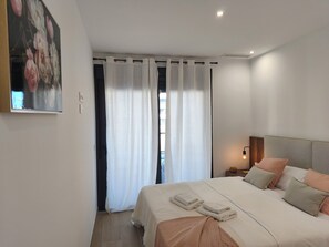 Ein Schlafzimmer mit einem Bett, Kissen, einem Nachttisch mit Lampe, einem Bild an der Wand und einem Fenster mit Vorhängen.