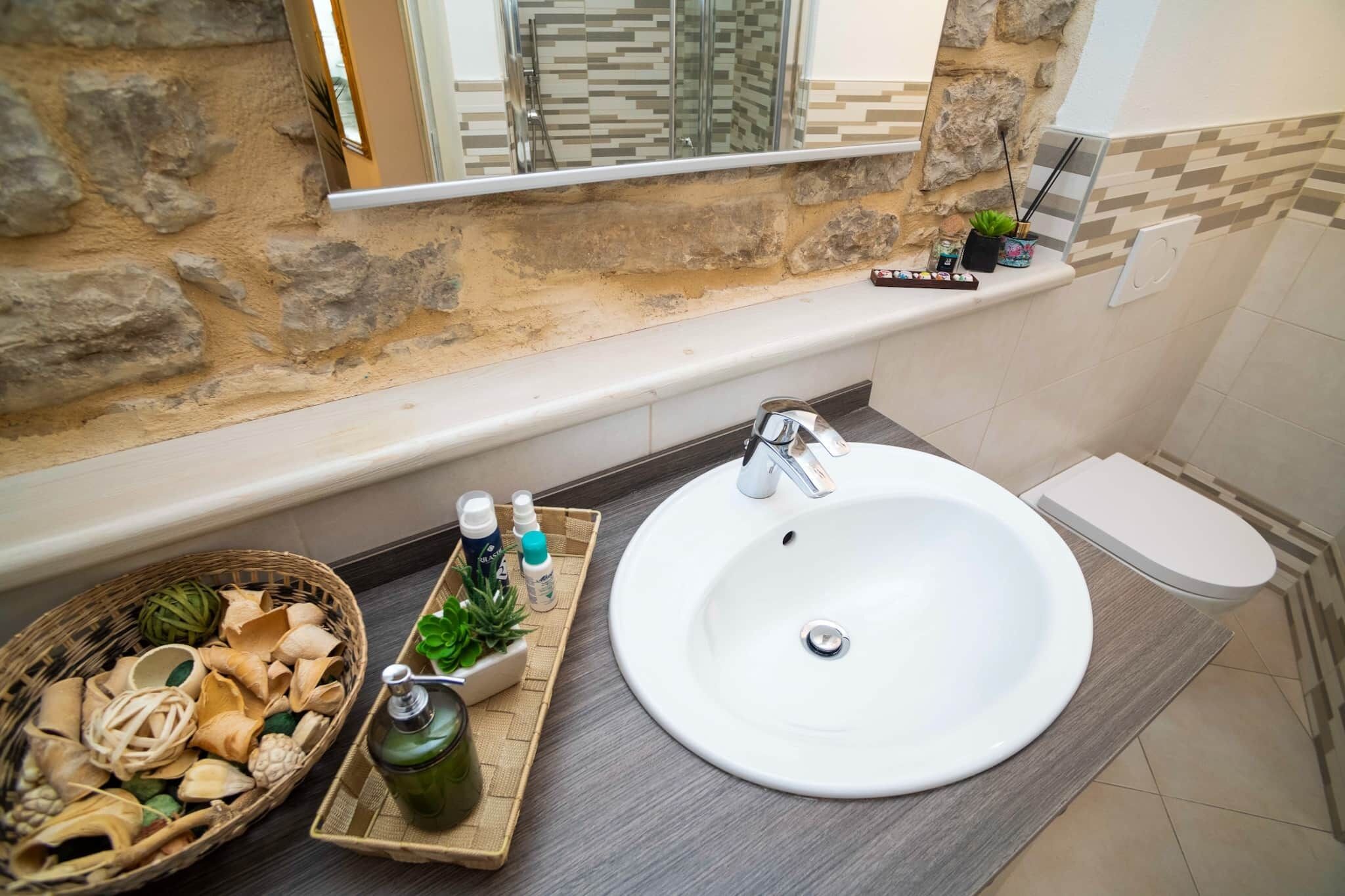 A bathroom with a stone wall, a large sink, a toilet, and a small shelf with toiletries.