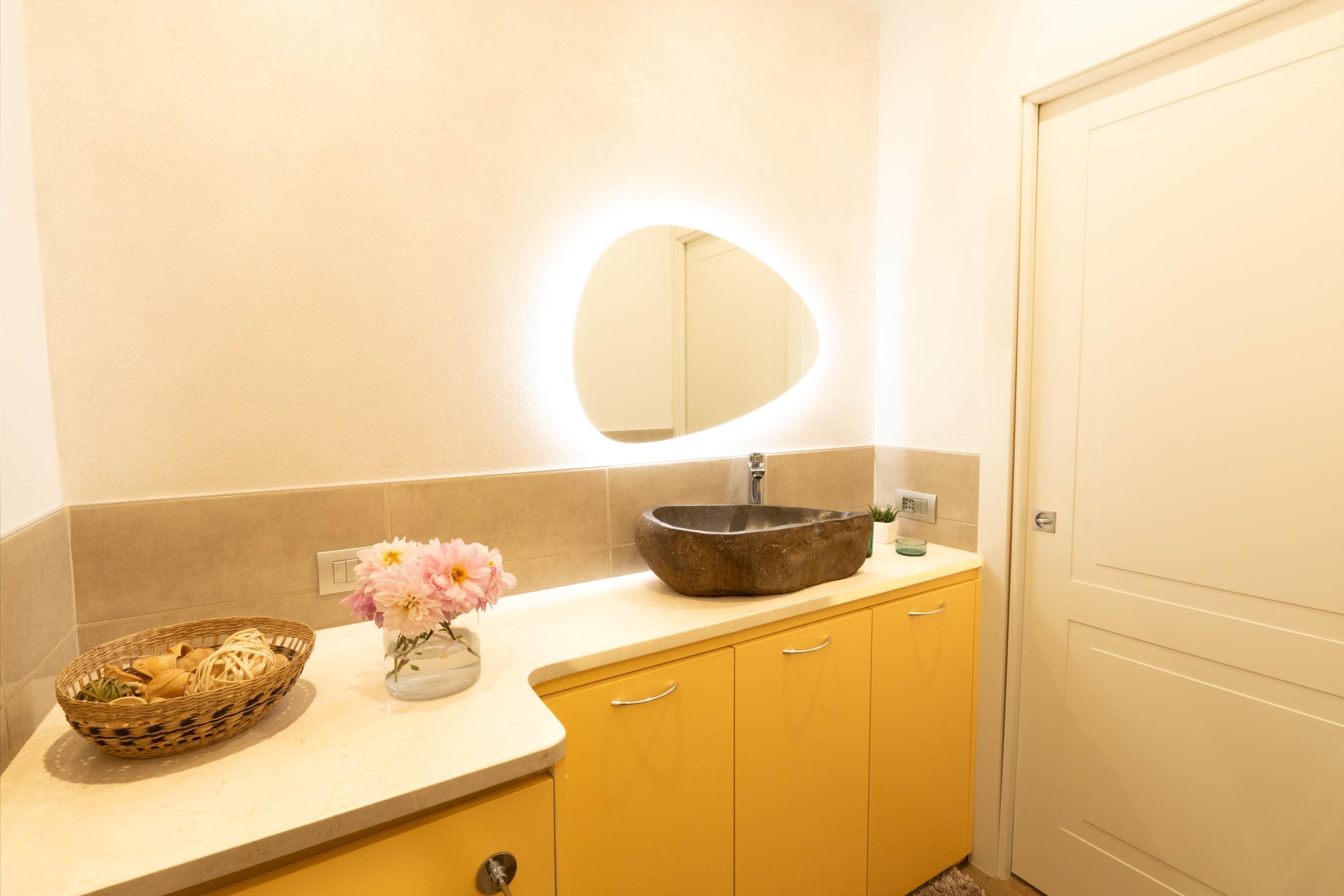 A bathroom with a round mirror, a stone sink, a wicker basket, and a vase with flowers.