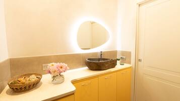 A bathroom with a round mirror, a stone sink, a wicker basket, and a vase with flowers.