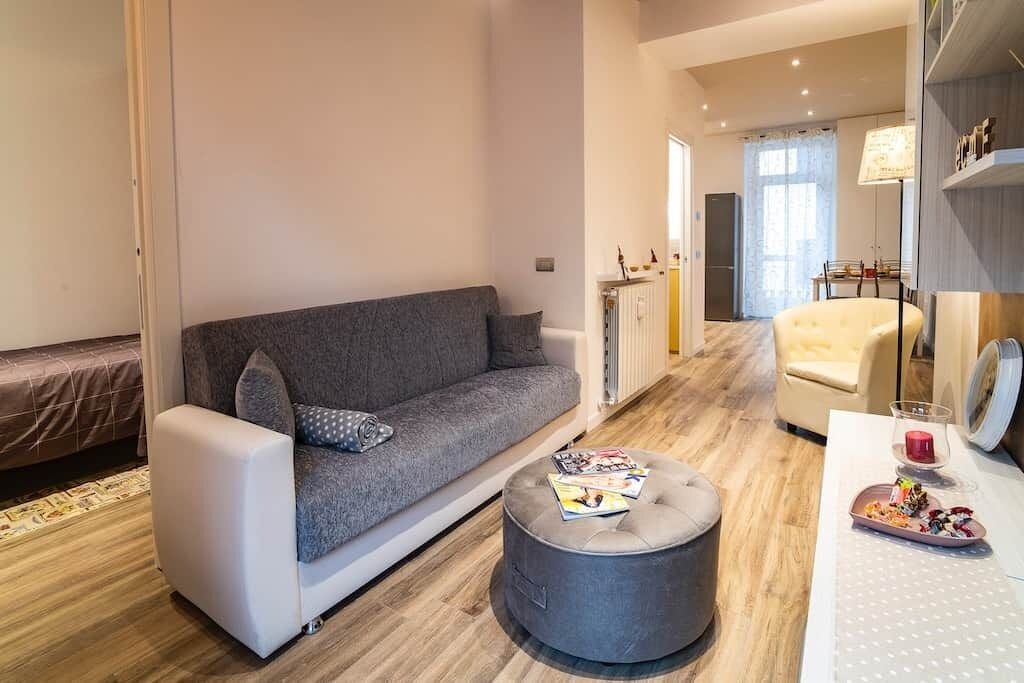 Un salon moderne avec un canapé gris, un pouf rond, une table à manger avec une lampe, et un coin cuisine à l’arrière-plan.