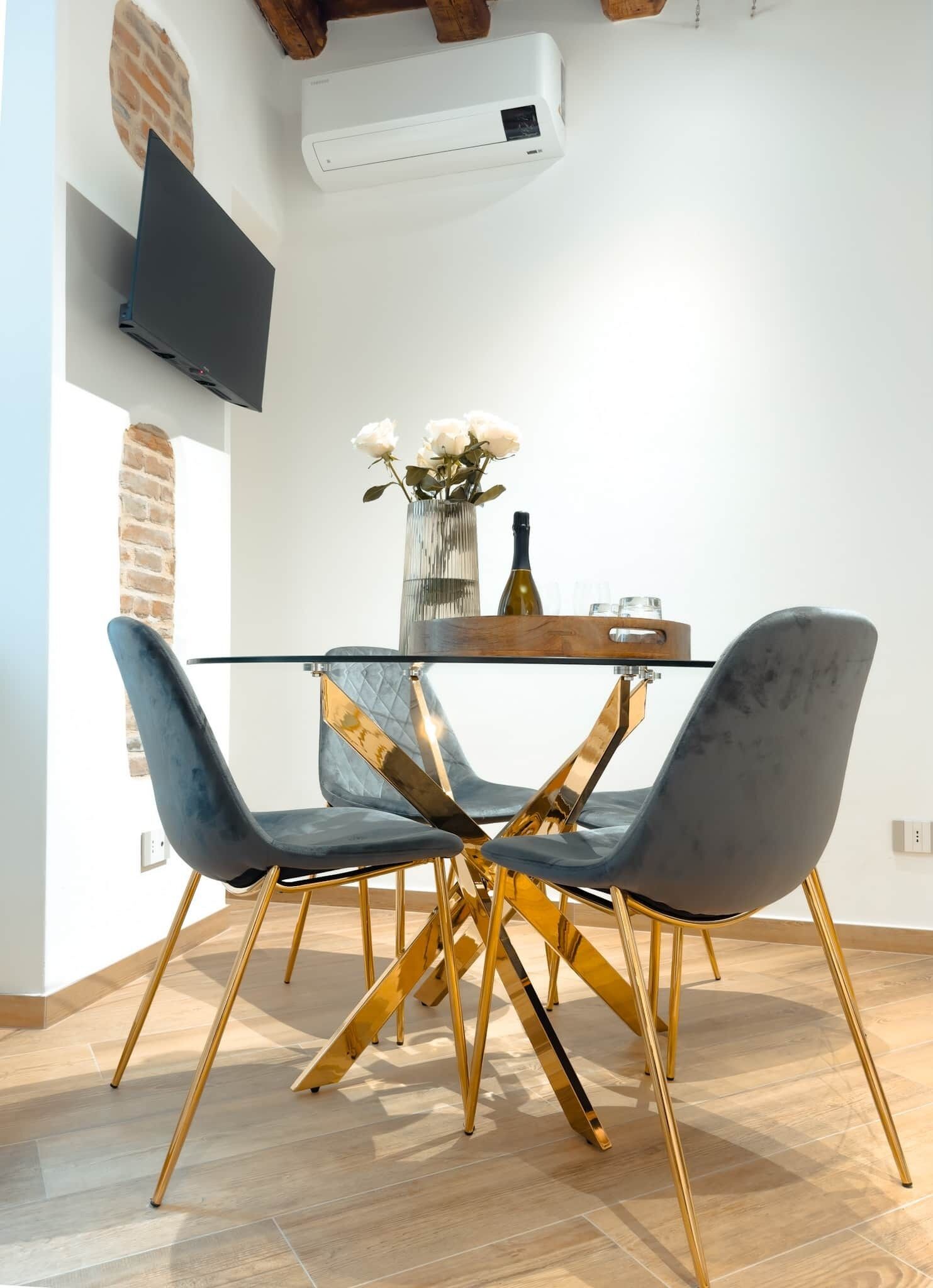 Un moderno comedor con una mesa de vidrio, dos sillas de terciopelo, un florero con flores y un televisor de pantalla plana montado en la pared.