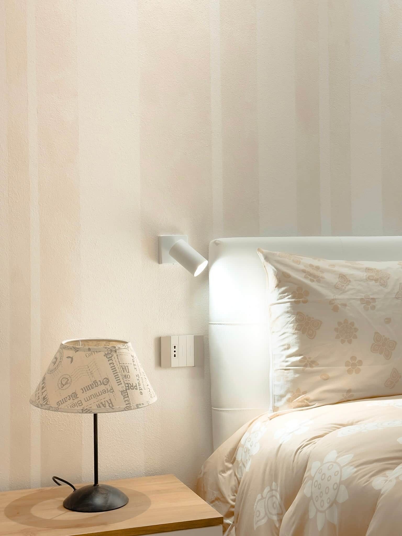 Ein ordentlich eingerichtetes Schlafzimmer mit Nachttischlampe, Wandleuchte und einem Bett mit floralem Bettbezug.