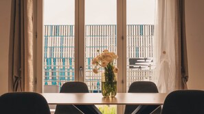 Une pièce comprenant une table, des chaises, un vase avec des fleurs et une fenêtre donnant sur un bâtiment aux motifs bleus et blancs.