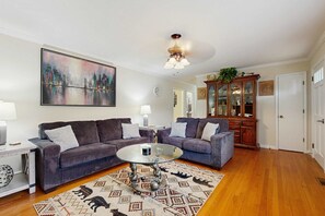 Ein Wohnzimmer mit zwei Sofas, einem Glastisch, einem Stadtbildgemälde, einem Holzschrank und einem Teppich mit Muster.