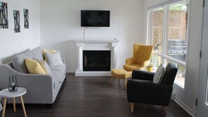 A living room with a fireplace, a sofa, a chair, and a TV mounted on the wall. - Waterfront & Spa | Le Petit Georges (Saint-Georges-de-Windsor)