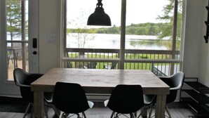 A dining area with a table and chairs, large windows with a view of a lake and greenery, and a hanging pendant light. - Waterfront & Spa | Le Petit Georges (Saint-Georges-de-Windsor)