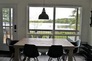 En matplats med ett bord och stolar, stora fönster med utsikt över en sjö och grönområden, samt en hängande taklampa.