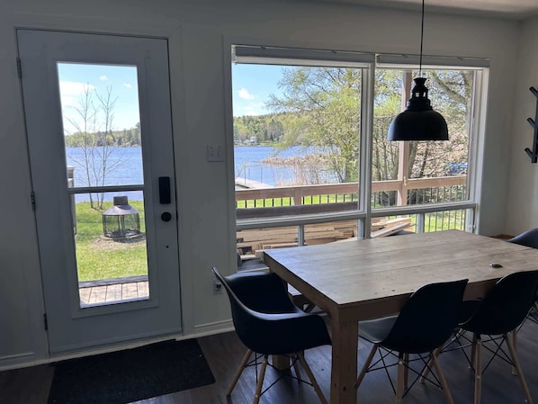 Comedor con mesa y sillas, ventanas amplias con vista a un lago y a una terraza, y una lámpara colgante.