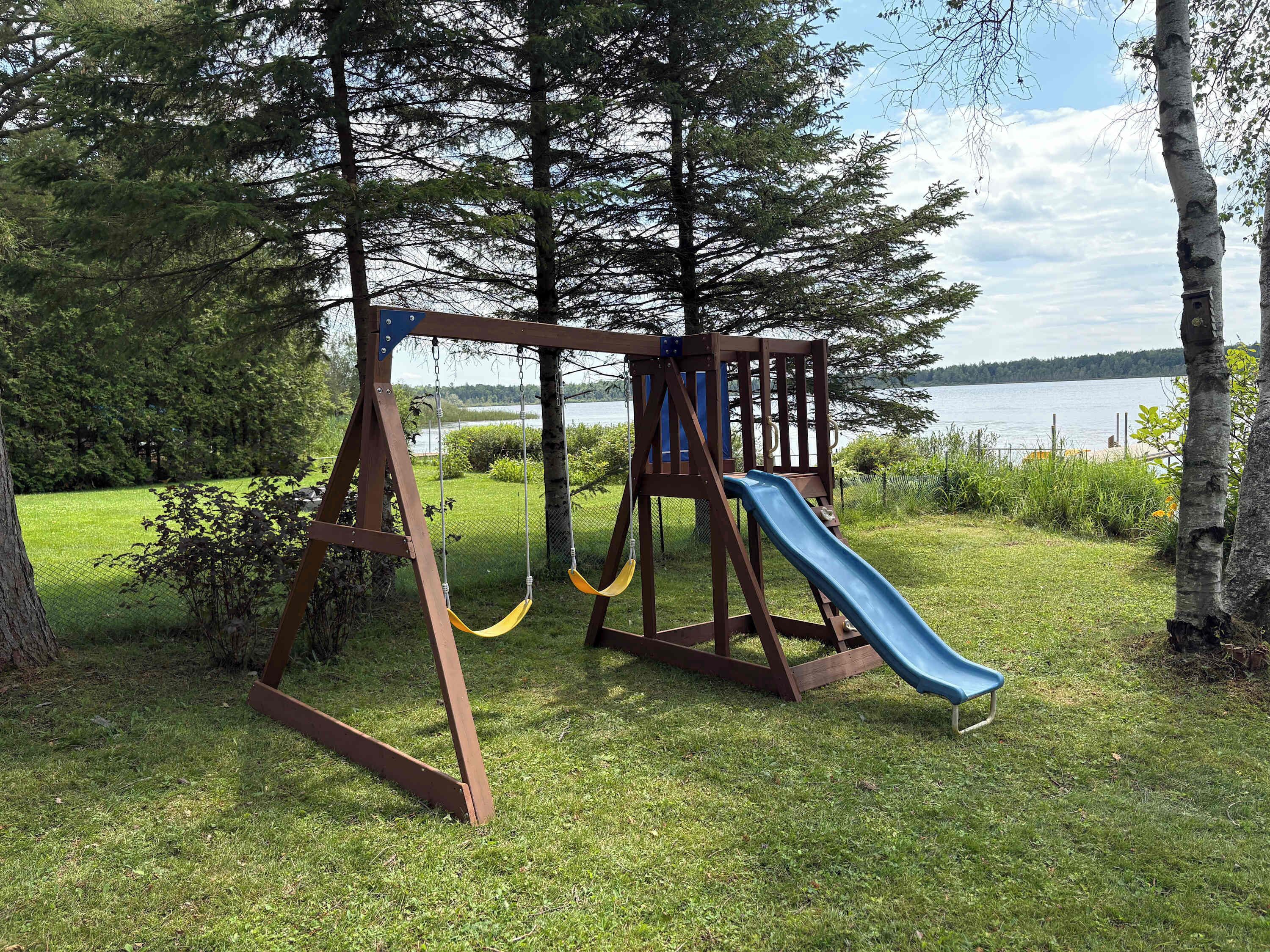Un parque infantil con tobogán y columpios, rodeado de árboles y vista a un lago.