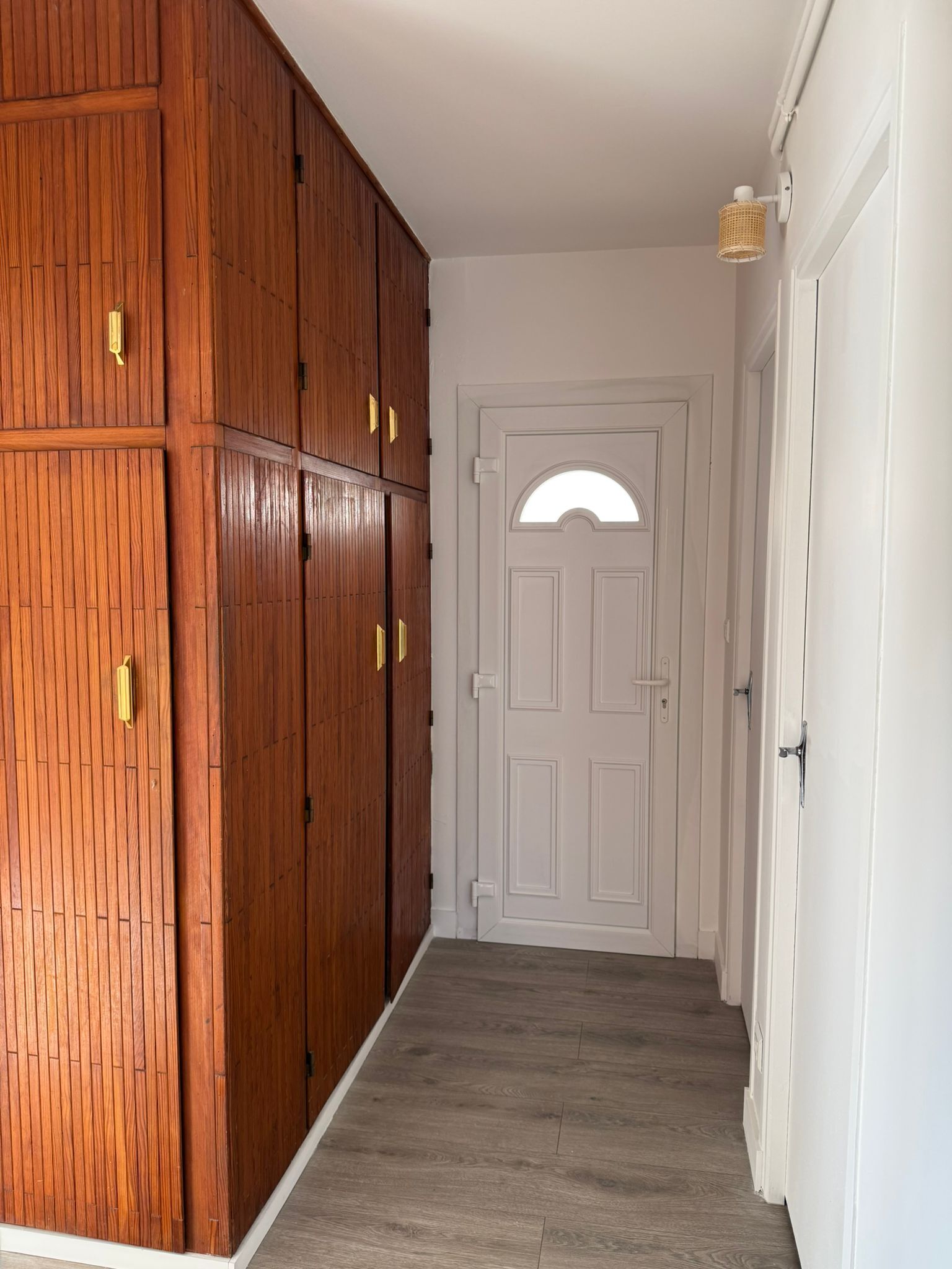 A hallway with a wooden wardrobe, a white door with an arched window, and a light fixture.