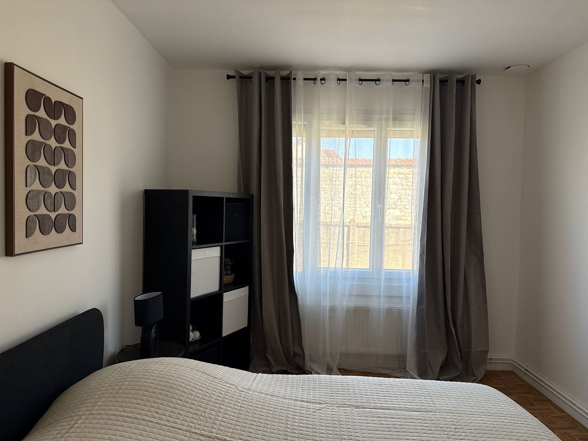 Une chambre à coucher avec un lit, une bibliothèque, une œuvre d’art accrochée au mur et une fenêtre avec des rideaux.