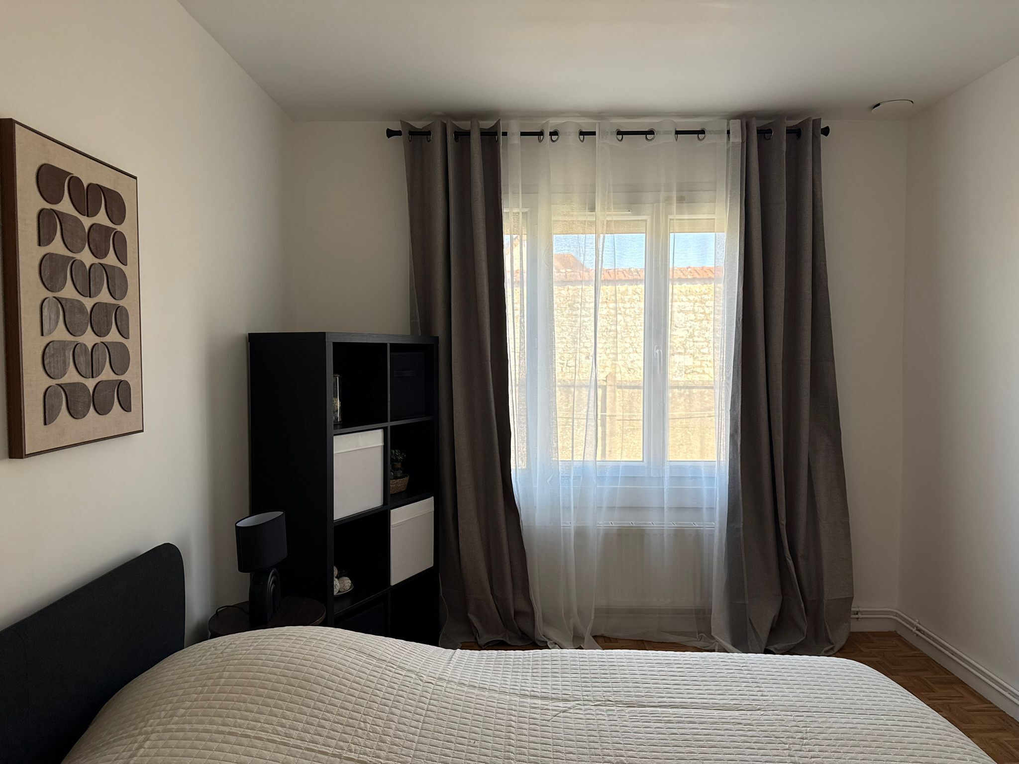 Un dormitorio con una cama, una estantería negra, una obra de arte en la pared y una ventana con cortinas translúcidas.
