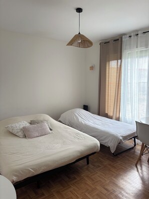 Una habitación con dos camas, piso de madera, lámpara colgante, una silla y un ventanal con cortinas.