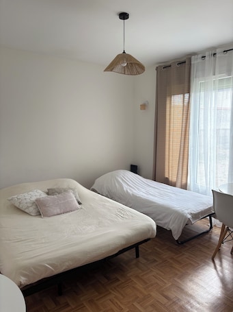 A room with two beds, a wooden floor, a hanging light fixture, a chair, and a window with curtains.. Maison du Potager & large garden center Vaudreuil