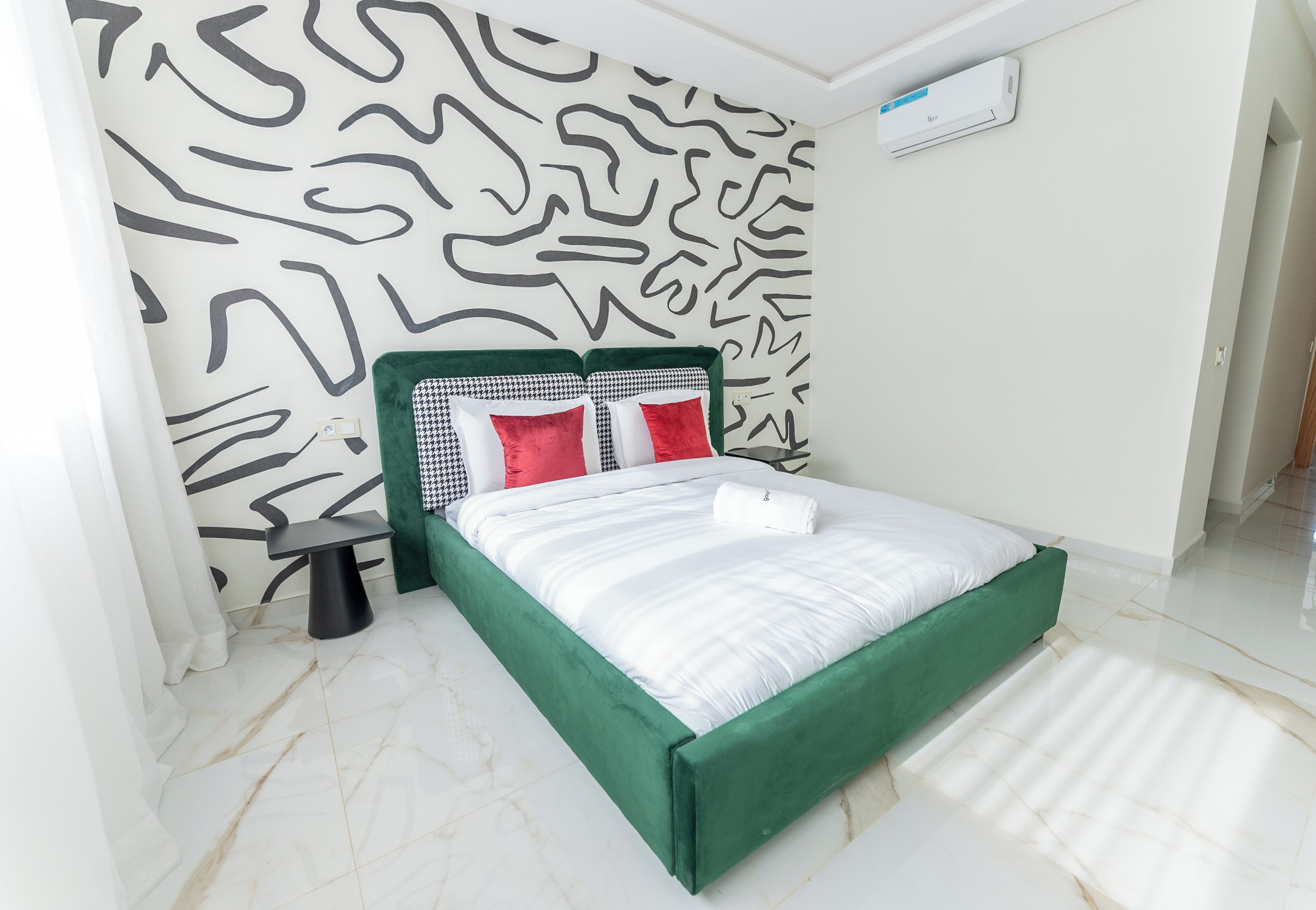 Uma cama com estrutura verde, roupa de cama branca e almofadas vermelhas. Há uma pequena mesa preta ao lado da cama. O quarto tem piso branco e parede estampada.