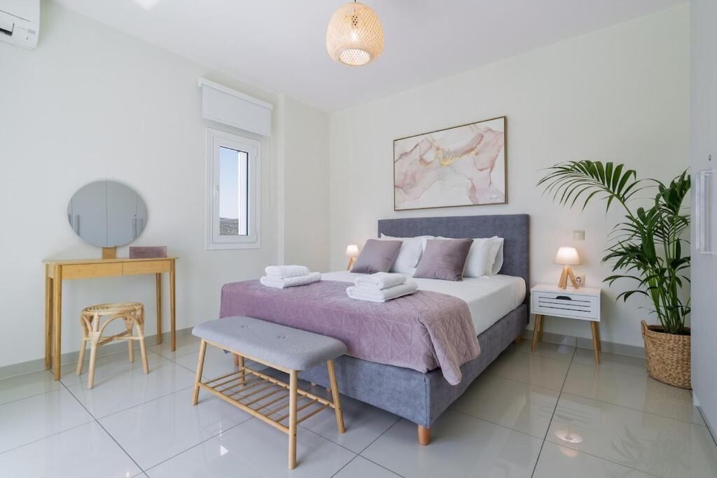 A bedroom with a bed, bedside tables, a bench, a round mirror, a window, and a potted plant.