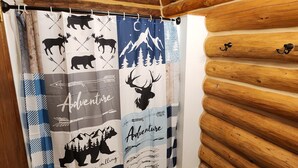 A bathroom with a wooden log wall, a shower curtain with wildlife and adventure-themed designs, and a checkered bath mat. - Private Log Cabin on 20 acres, Across from Custer SP, Close to Mt Rushmore (Custer)