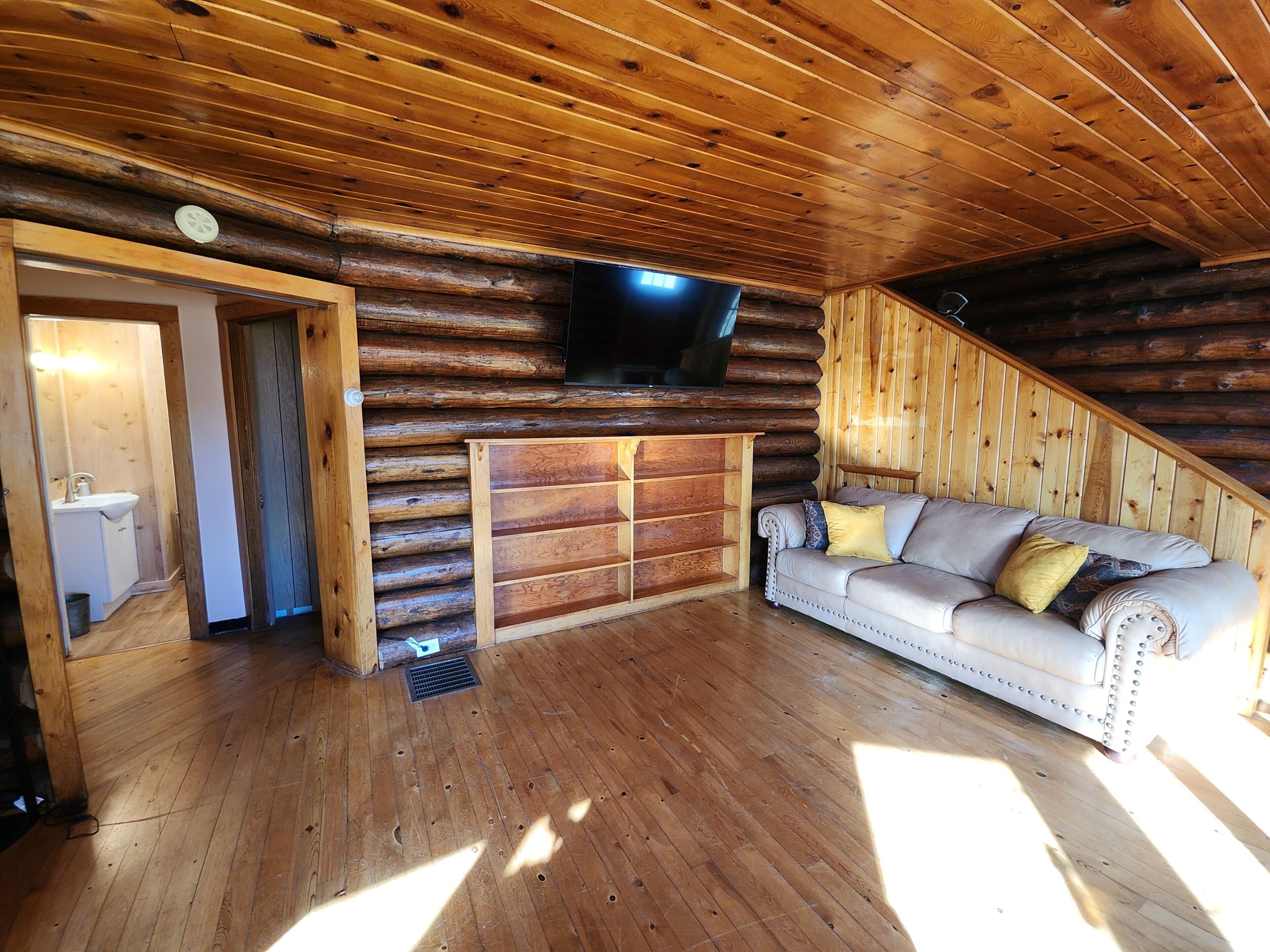 Interior de una cabaña de troncos con suelo de madera, un sofá, un televisor montado en la pared y un baño pequeño visible a través de una puerta abierta.