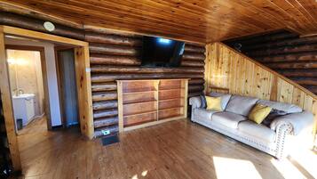 Interior de una cabaña de troncos con suelo de madera, un sofá, un televisor montado en la pared y un baño pequeño visible a través de una puerta abierta.
