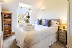 A bedroom with a large bed, a bedside table, a lamp, a bookshelf, and a view of the outside. - Mill Village 43, Dragonfly - Spa & Swimming Pool Access, Watersports & Pet Friendly, Sleeps 6 (Somerford Keynes)