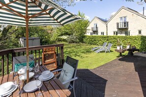 A wooden deck with a table set for dining, a lounge chair, and a striped umbrella. - Mill Village 43, Dragonfly - Spa & Swimming Pool Access, Watersports & Pet Friendly, Sleeps 6 (Somerford Keynes)